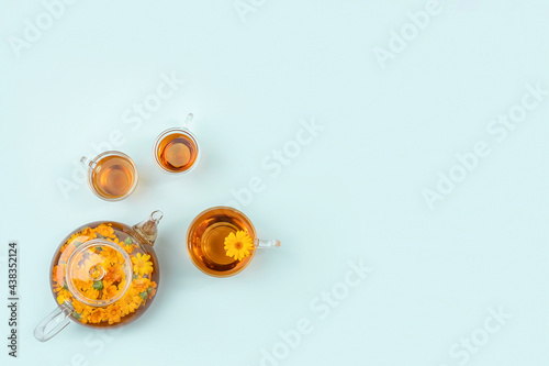 Cups of herbal tea, transparent teapot with calendula flowers on blue background. Calming drink concept. Copy space Flat lay Top view