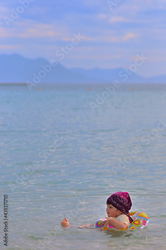 A little girl in a bandana floats on an inflatable ring alone in the sea against a background of blurry mountains and clouds. Side view, soft lilac background for copy space. Vietnam, Nha Trang.