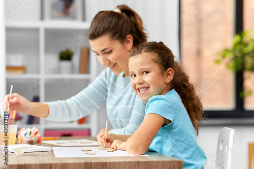 family, hobby and leisure concept - happy smiling mother spending time with her little daughter drawing or painting wooden chipboard cutouts with colors at home