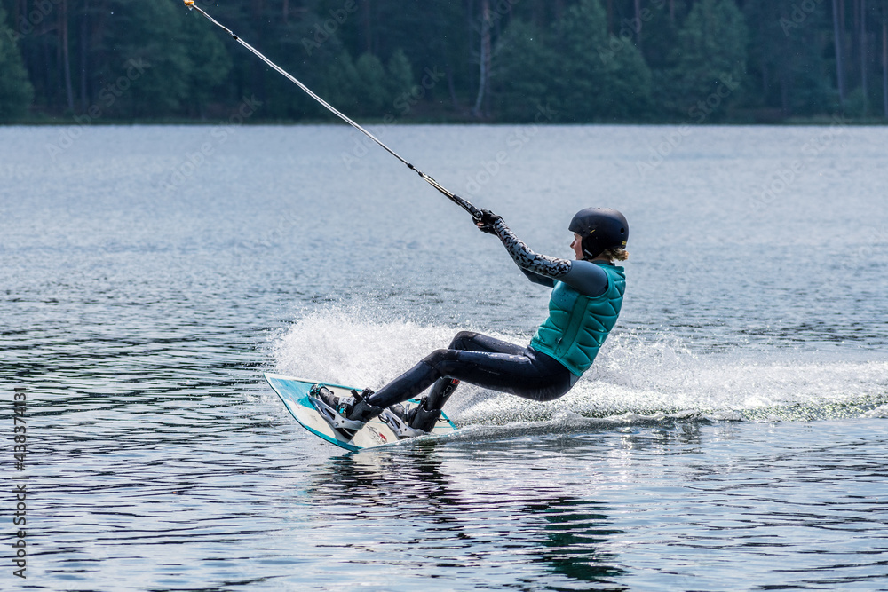 Naklejka premium Girl with helmet and life vest rides a wakeboard on the lake with forest on background, wakeboarding, water sport in summer 
