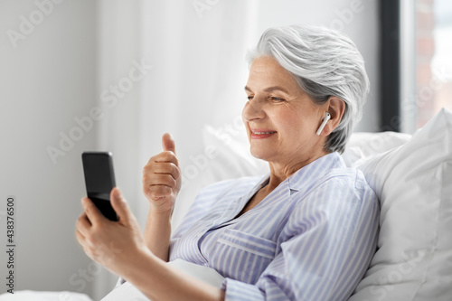 technology, old age and people concept - senior woman with smartphone and wireless earphones having video call in bed at home bedroom