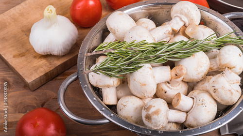 vegetarian cuisine - champignons in a colander with rosemary branches on the table with ingredients for cooking a vegan dish, vegetables, tomatoes, garlic and onions
