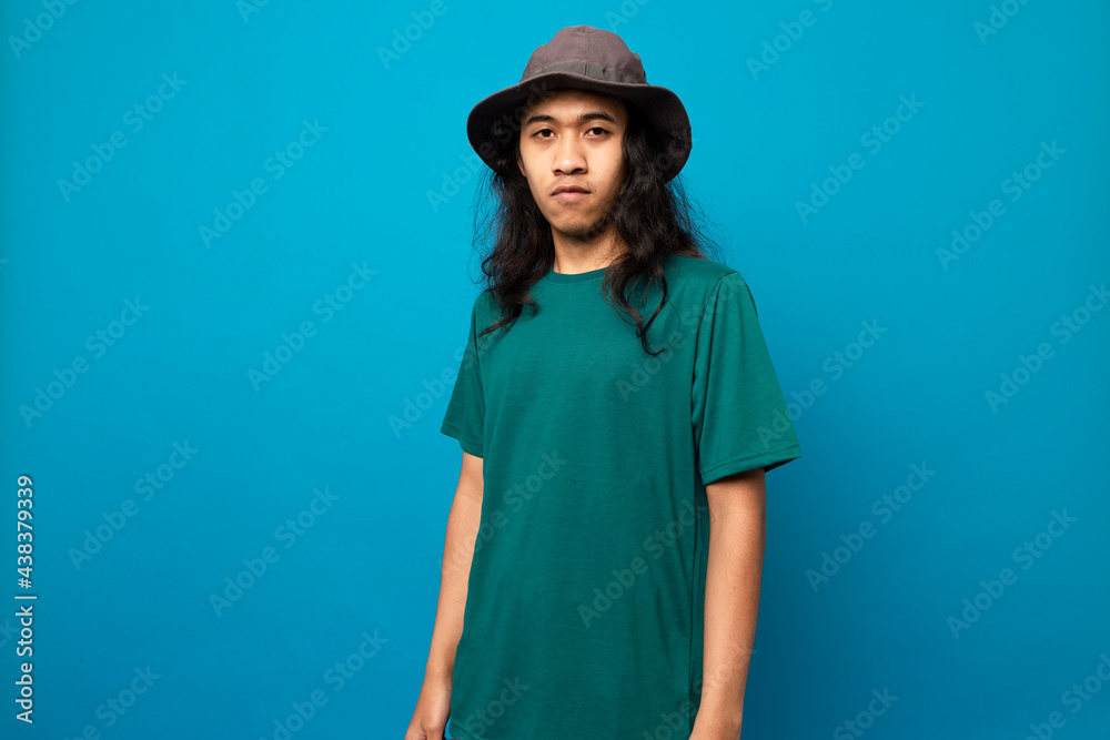 young man with long hair wearing a hat with a serious face isolated on blue background