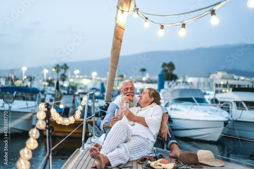 Senior couple drinking champagne and eating fruits on sailboat during summer vacation - Focus on faces