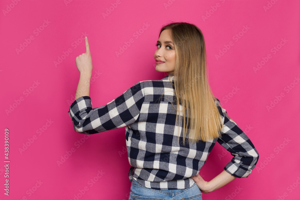 Young Woman In Lumberjack Shirt Is Pointing Up And Looking At Camera Over The Shoulder. Rear View. Young Woman In Lumberjack Shirt Is Pointing Up And Looking At Camera Over The Shoulder. Rear View.