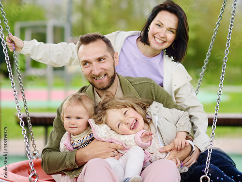 Merry family with two children having fun on outdoor playground. Young parents rides daughters on swing. Spring/summer/autumn active leisure for family with kids.