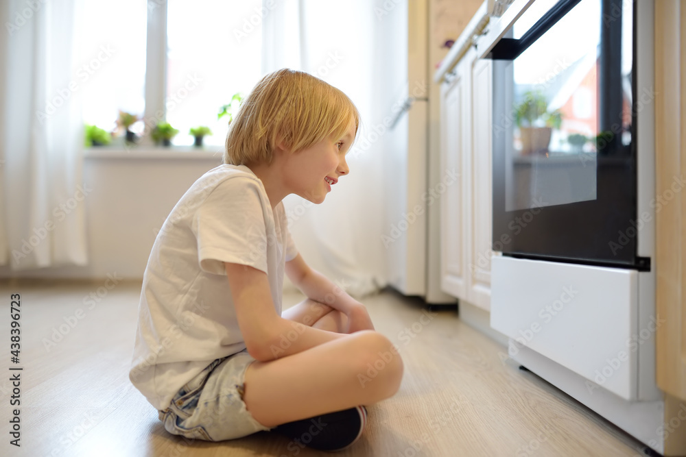 Glad boy sitting on the floor near kitchen stove and waiting for a pie or other baked goods to be prepared. Children love food prepared by their mother. Domestic cuisine Glad boy sitting on the floor near kitchen stove and waiting for a pie or other baked goods to be prepared. Children love food prepared by their mother. Domestic cuisine