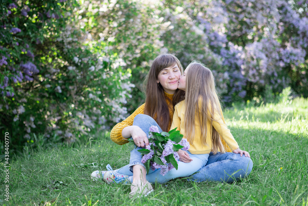 Fototapeta premium Mom and daughter laughing and hugging sitting on a blanket in the park in a clearing. The concept of a happy family, friendship, and love. Outdoor activities