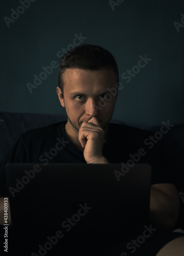 Close up dark portrait of young adult man thinking near laptop watching in camera. Male typing uses computer, holds hand near face, leans on fingers. Dark natural light atmosphere, gloom from window