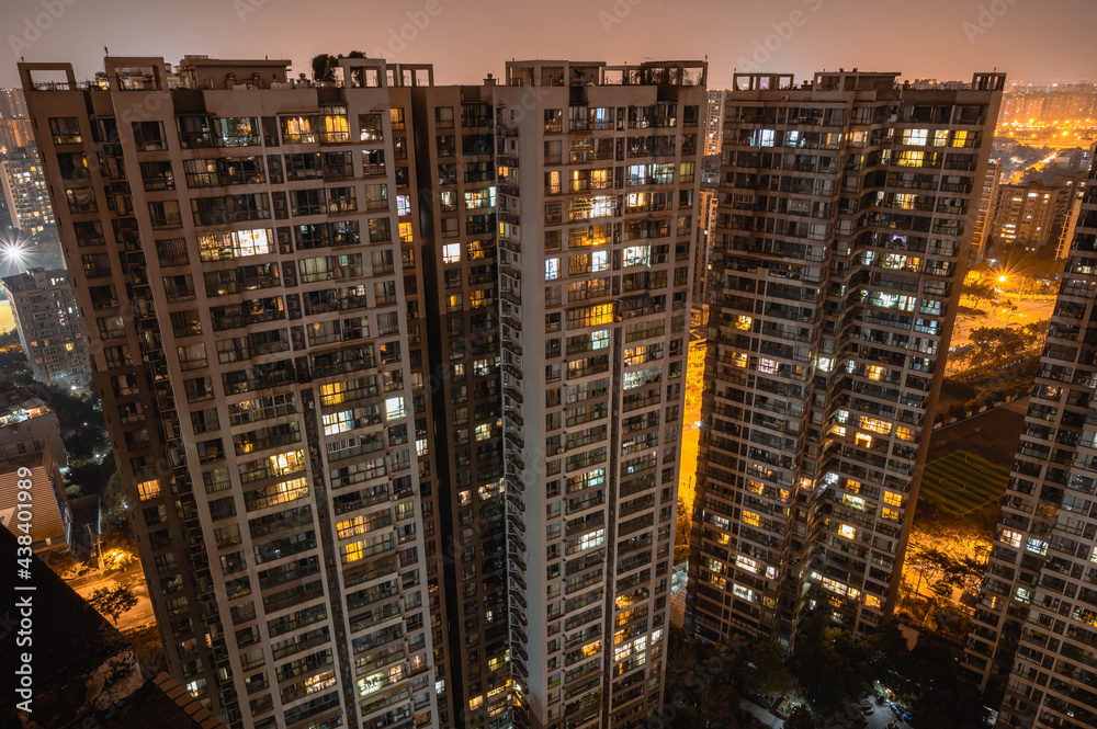 Long exposure night aerial view on residential buildings complex compound. Tall skyscrapers thousands of apartments light illuminates from windows. Small square park inside community. City development