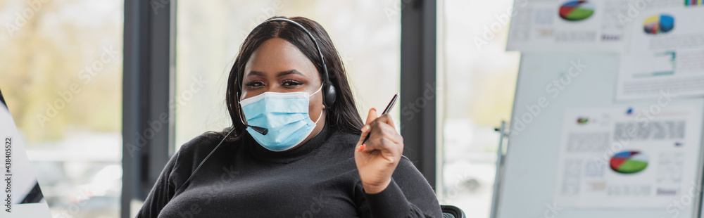 african american plus size operator in headset and medical mask holding pen in modern office, banner. african american plus size operator in headset and medical mask holding pen in modern office, banner.
