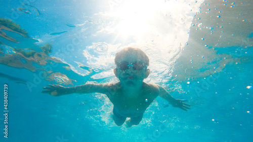 Underwater photo of a kid with goggles swimming into a pool with bubbles around and blue sky behind. Active healthy lifestyle, water sport activity and lessons on summer holidays vacation.