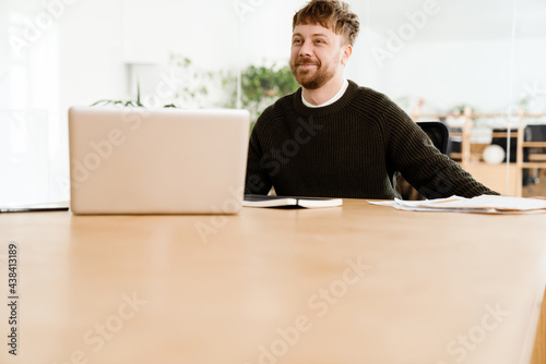 Young ginger man working with laptop while sitting at desk in office