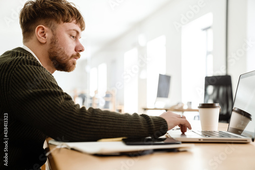 Young ginger man working with laptop while sitting at table in office