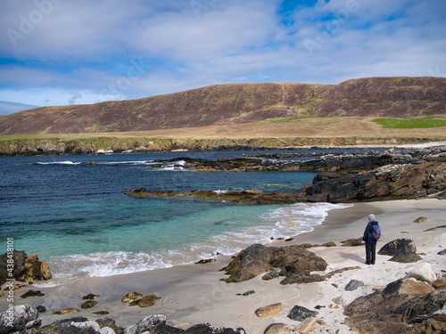 With turquoise, clear water and white sand, a single female walker looks to the sea on a beach at Collaster on the west coast of the island of Unst in Shetland, UK - taken on a calm sunny day