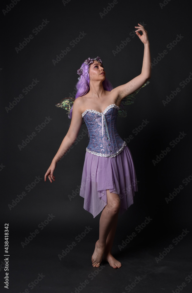 Full length portrait of a purple haired  girl wearing fantasy corset dress with fairy wings and flower crown.  Standing pose against a dark studio background.