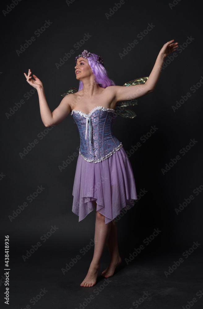 Naklejka premium Full length portrait of a purple haired girl wearing fantasy corset dress with fairy wings and flower crown. Standing pose against a dark studio background.