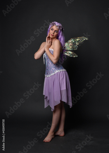 Full length portrait of a purple haired girl wearing fantasy corset dress with fairy wings and flower crown. Standing pose against a dark studio background.