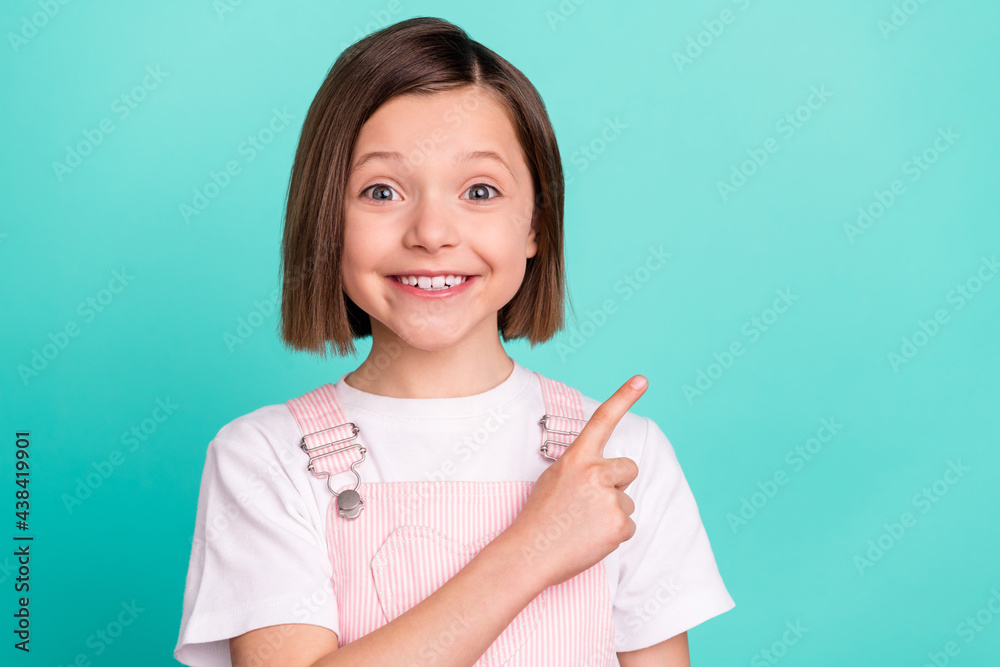 Photo of adorable pretty schoolgirl dressed pink overall pointing finger empty space smiling isolated turquoise color background Photo of adorable pretty schoolgirl dressed pink overall pointing finger empty space smiling isolated turquoise color background