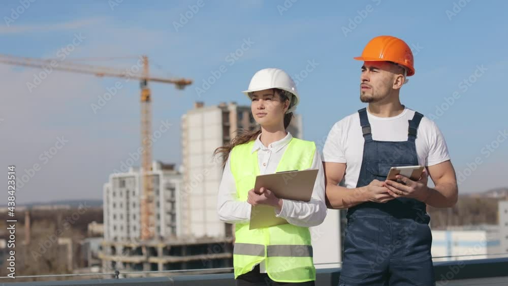 Two serious engineers are standing and looking away. They are wearing work clothes and a hard hat. They are on the roof of a business center. Construction in the background. Nice sunny day. 4K Two serious engineers are standing and looking away. They are wearing work clothes and a hard hat. They are on the roof of a business center. Construction in the background. Nice sunny day. 4K