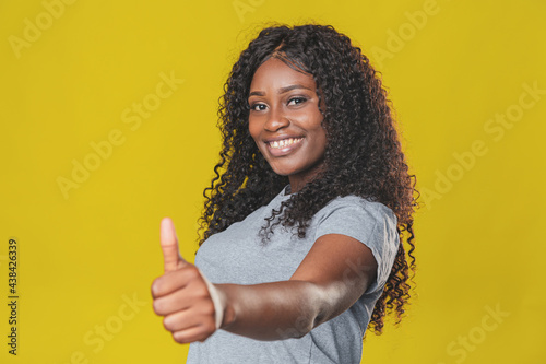 Beautiful african american girl with afro hairstyle in a gray t-shirt smiles and shows thumbs up on a yellow background © Nana_studio