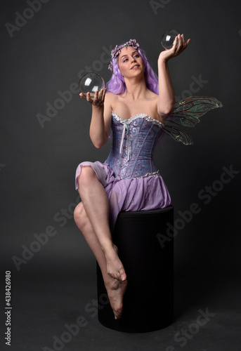 Full length portrait of a purple haired girl wearing fantasy corset dress with fairy wings and flower crown. Seated pose against a dark studio background.