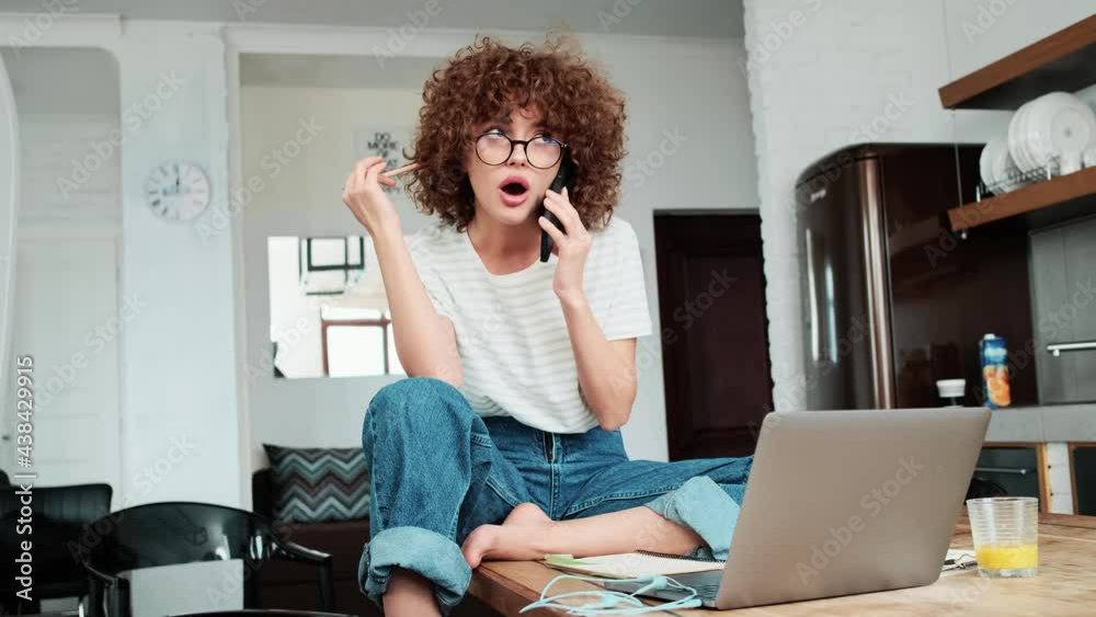 A surprised curly-haired girl in an eyeglasses talking at the phone and looking at the laptop at home A surprised curly-haired girl in an eyeglasses talking at the phone and looking at the laptop at home