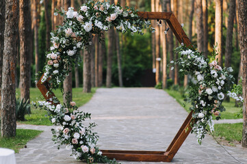 A beautiful wooden wedding arch in the form of a hexagon, decorated with natural flowers, stands on the road against the background of a forest with pine trees.