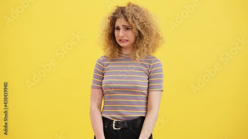A confused curly blonde woman is looking at something disgusting standing isolated over yellow wall in the studio photo