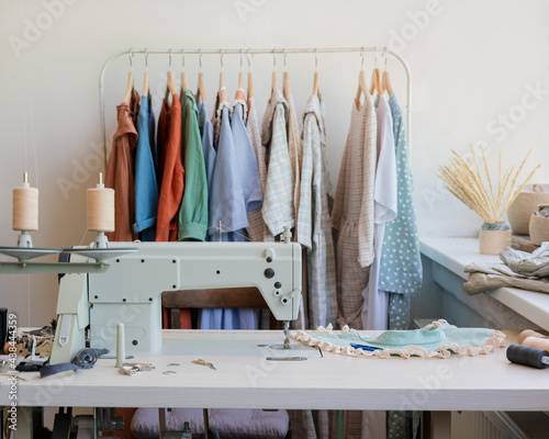 Cozy interior of fashion design studio with professional sewing machine and various sewing accessories on table, workplace of dressmaker or fashion designer with exclusive trendy clothes on hangers