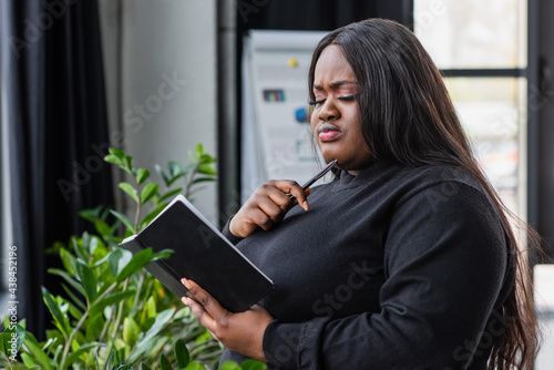 pensive african american plus size businesswoman holding notebook in office.