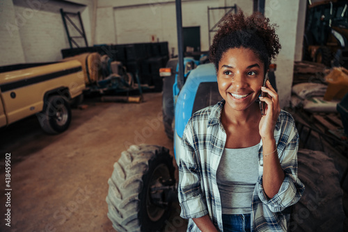 Mixed race female chatting on cellular device to colleagues leaning on tractor in stable