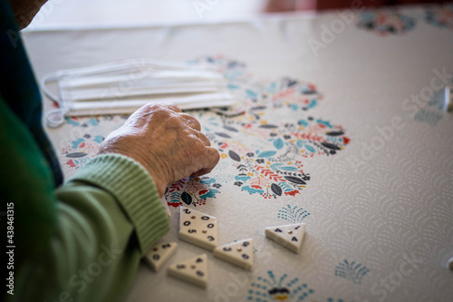 Main d'une femme âgée qui joue à un jeu de société sur une table napée, avec un masque dans le fond photo