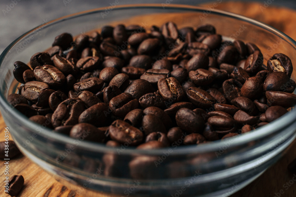 Fototapeta premium Coffee beans in a transparent bowl over a wood tablet with copy space. Dark background of coffee beans.
