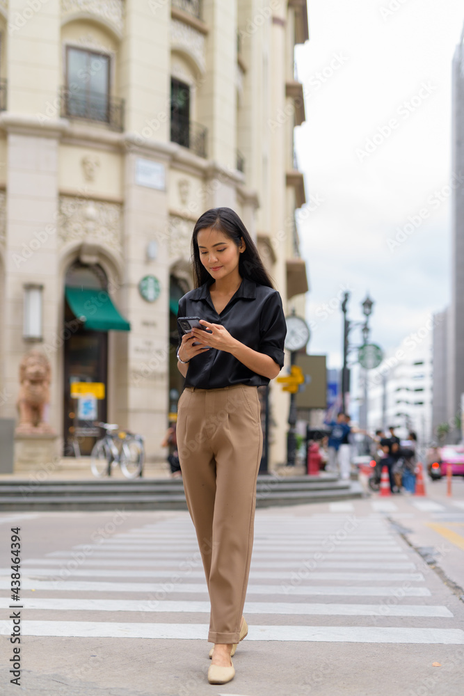 Beautiful Asian businesswoman walking outdoors in city street using mobile phone and texting