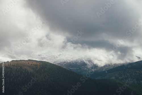 Atmospheric autumn landscape with forest mountains in snow in low clouds in overcast weather. Awesome scenery with gray rain clouds above snowy hills and mountains in autumn colors. Grainy clouds.