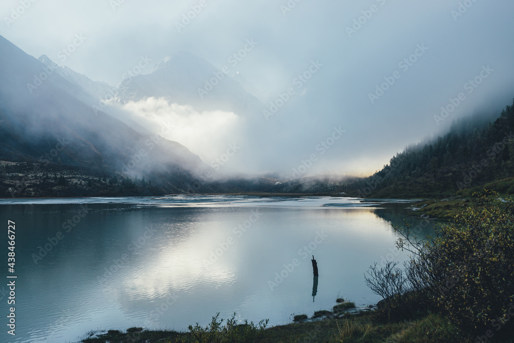 Atmospheric alpine landscape with mountain lake and high snow-covered mountain in dense low clouds. Beautiful low lighting scenery with pointy peak in thick fog and golden sunshine reflection in lake.