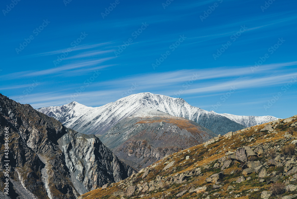 Colorful alpine landscape with great mountain in autumn colors with snow on peak in sunshine under cirrus clouds in blue sky. Picturesque autumn scenery with sunlit rocks and snow-covered mountain top