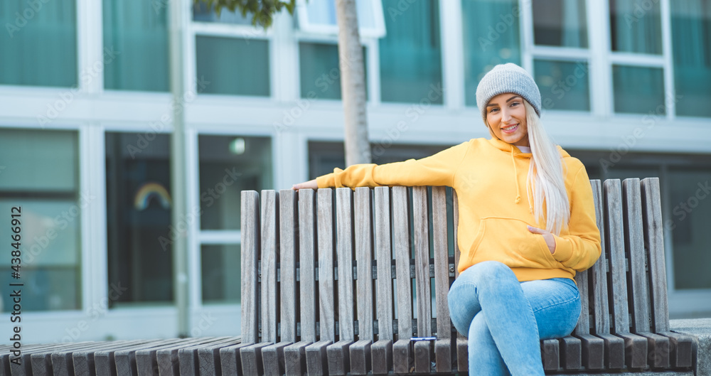Smiling full length beautiful young woman sitting on bench. Outdoors daylight. Pretty smile girl wearing casual clothing.