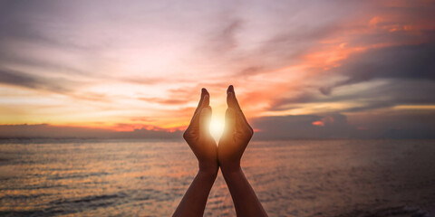 June summer solstice sun concept with silhouette of happy young woman's hands relaxing, meditating and holding sunset against warm golden hour sky on the beach with natural ocean or sea background