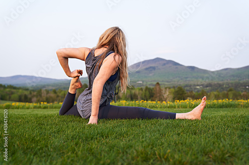 blonde yoga instructor stretches her legs in a split under bright sky with mountains in the background
