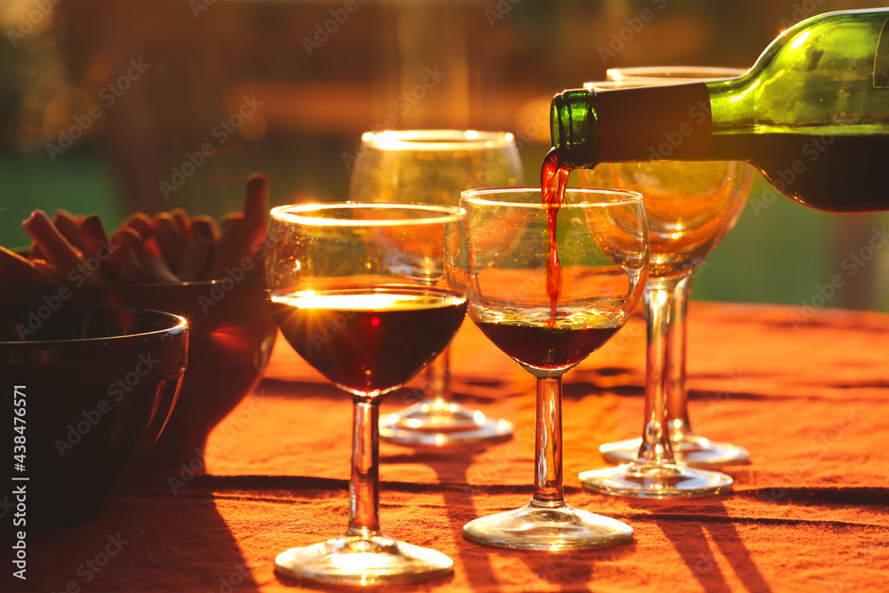 Fototapeta premium Pouring red wine from bottle into glasses on table with snacks background during the sunset