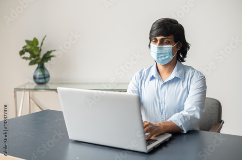 Portrait of focused young indian man wearing surgical medical mask and smart casual blue shirt sitting at the desk, using laptop and protects himself from virus diseases during office work