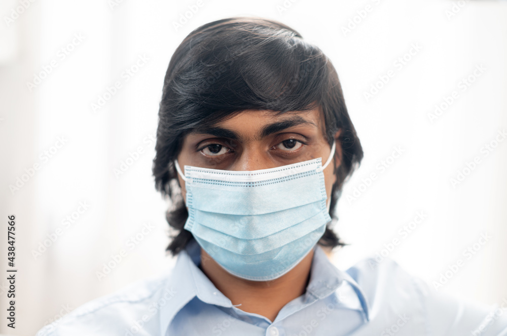 Close-up portrait of serious young indian man wearing surgical medical mask and smart casual blue shirt, eastern man protects himself from virus diseases, preventing spread of corona virus