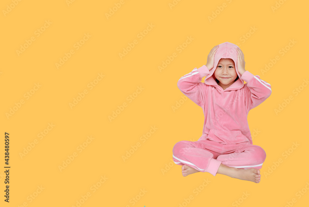 Happy little Asian child girl in pink tracksuit or sport cloth with hands touching hood on head sitting isolated on orange background with copy space. Happy little Asian child girl in pink tracksuit or sport cloth with hands touching hood on head sitting isolated on orange background with copy space.