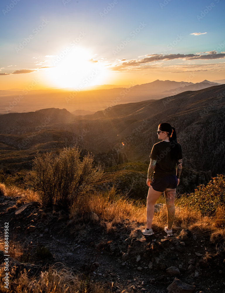 View of a young woman standing in the sunset looking out at mountains. She is a brunette with tattoo sleeves.