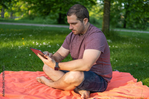 Obraz na płótnie Photo of a young and attractive man sitting in the park on a towel enjoying a sunny day outside in the nature and checking his tablet. Electronic device, technology w salonie