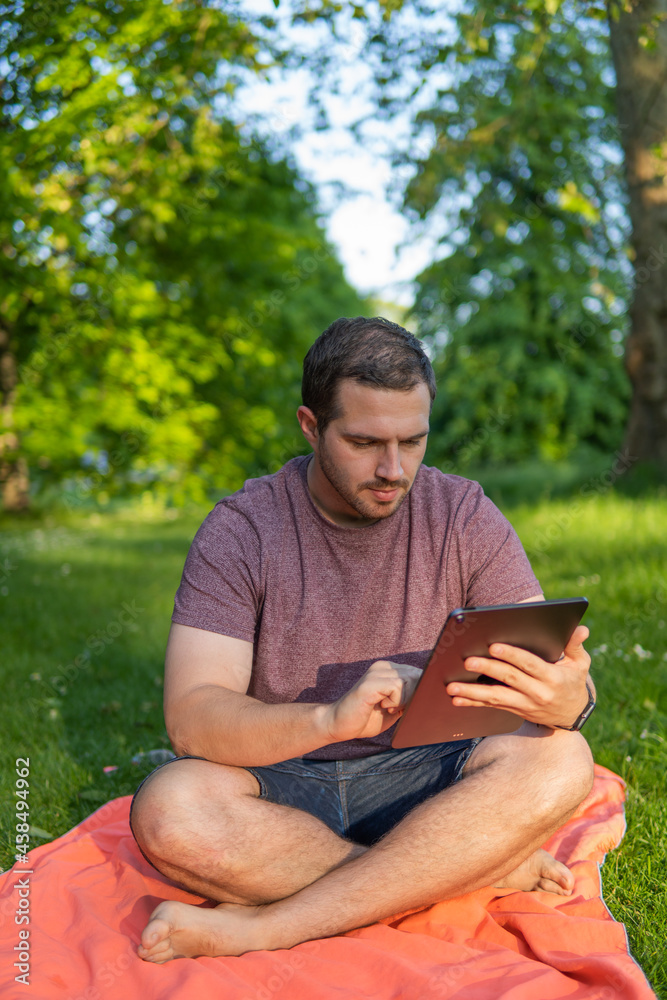 Photo of a young and attractive man sitting in the park on a towel enjoying a sunny day outside in the nature and checking his tablet. Electronic device, technology