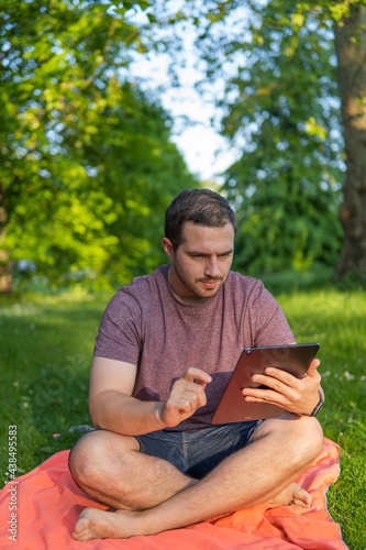 Photo of a young and attractive man sitting in the park on a towel enjoying a sunny day outside in the nature and checking his tablet. Electronic device  technology