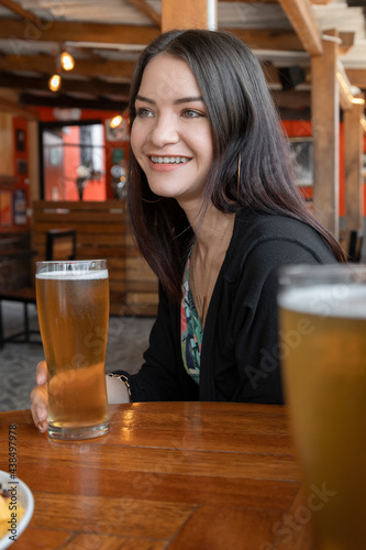 young woman with long straight hair sitting at a bar table with wooden decoration  holding a crystal glass with beer while smiling